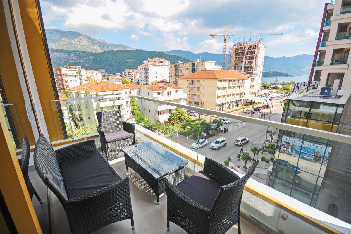 A balcony with wicker furniture and a glass table overlooking a cityscape with buildings, a street, and mountains in the background.