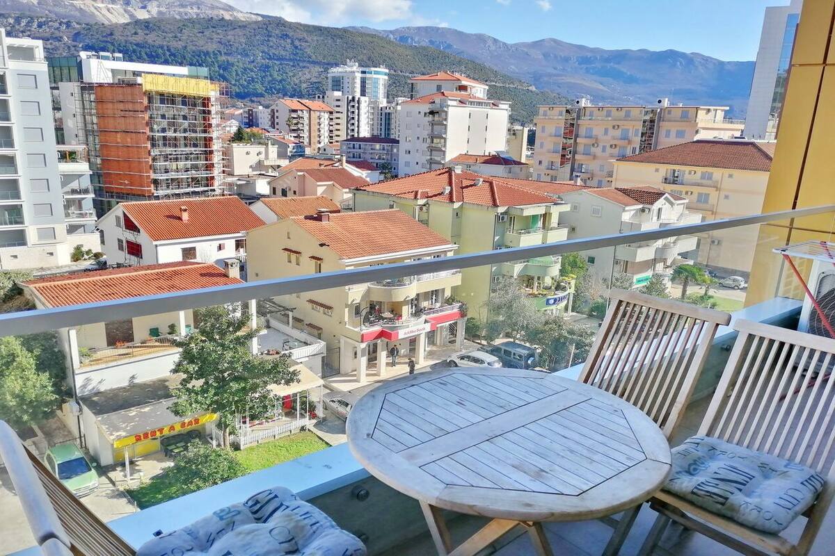 A balcony with a round table and chairs, overlooking a cityscape with buildings and mountains in the background.