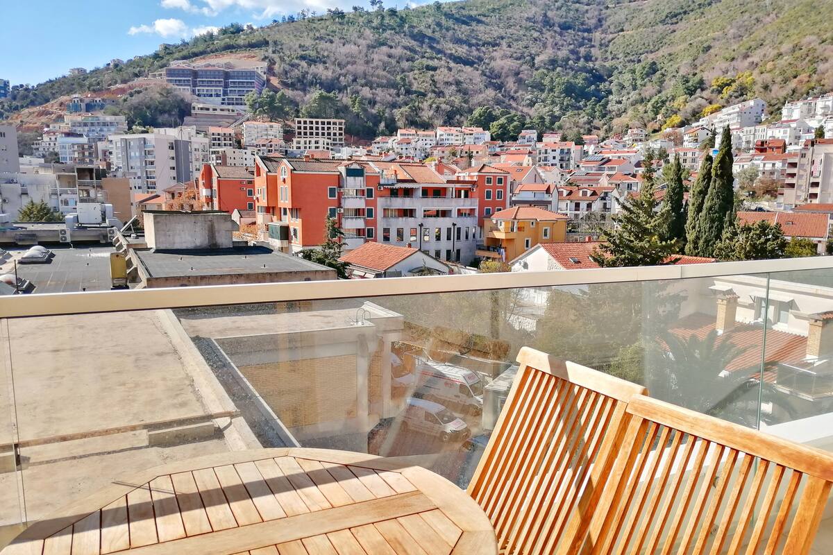 A rooftop terrace with a wooden table and chairs, overlooking a hillside town with buildings and trees.