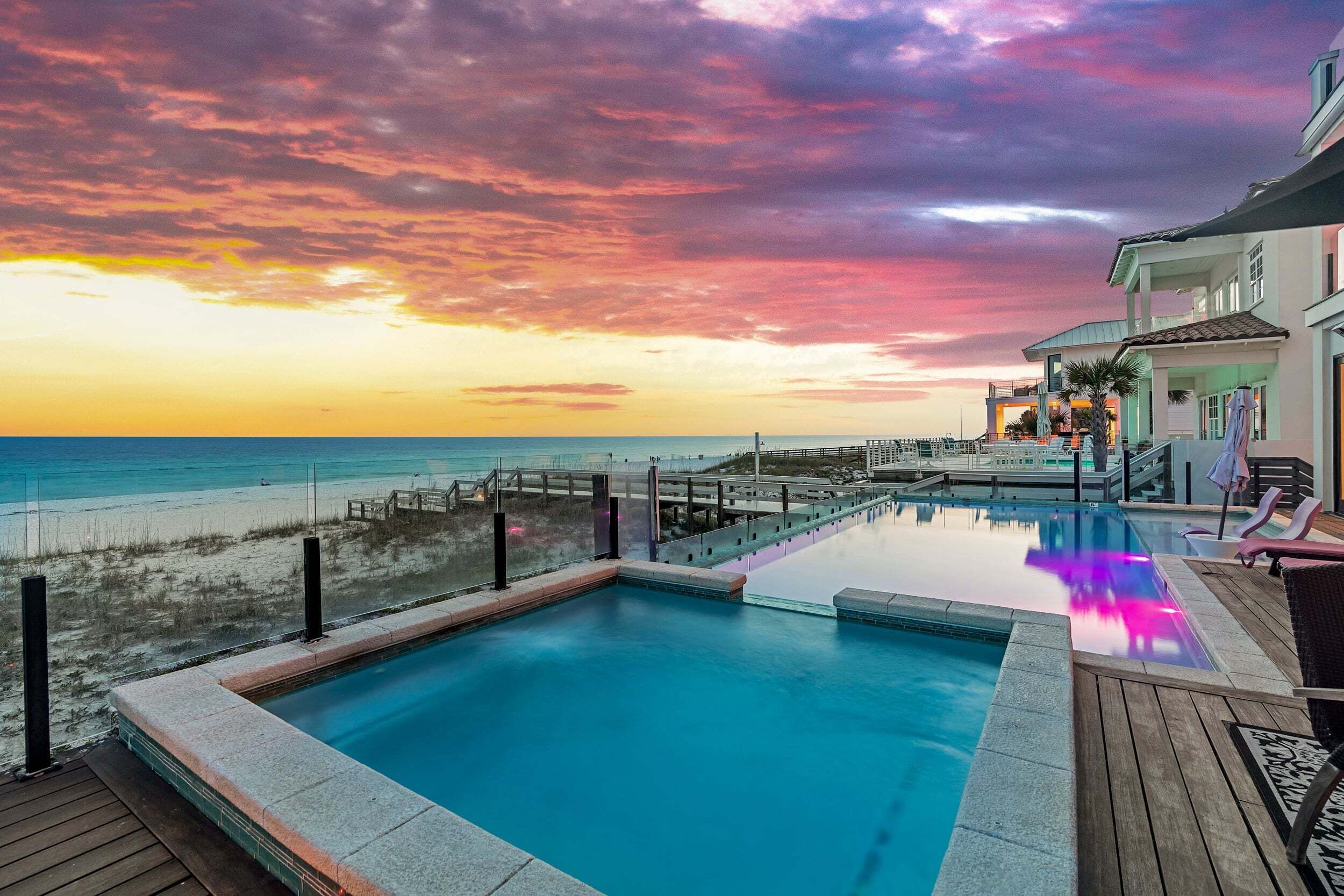 A pool with a wooden deck, a beach view, and a sunset sky.