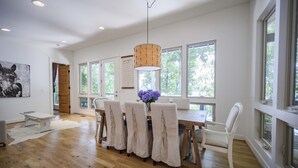 Un espace repas comprenant une table en bois, des chaises blanches, un luminaire suspendu et de grandes fenêtres donnant sur la forêt.
