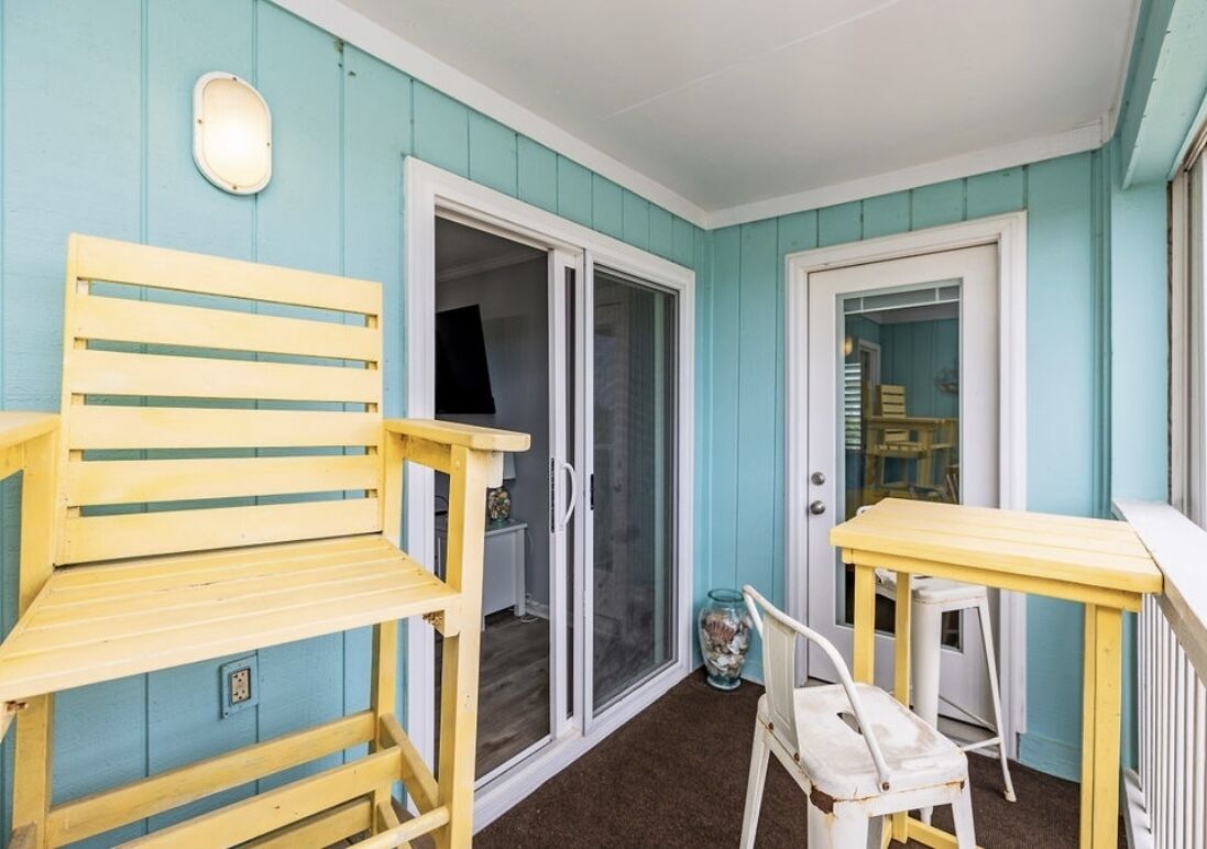 Un petit porche fermé, avec un banc en bois et une table, une porte-fenêtre coulissante et une petite chaise blanche.