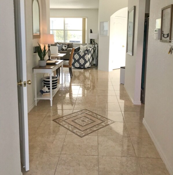 A hallway with a tiled floor, a dining area with a table and chairs, and a living room with a sofa and a patterned rug.