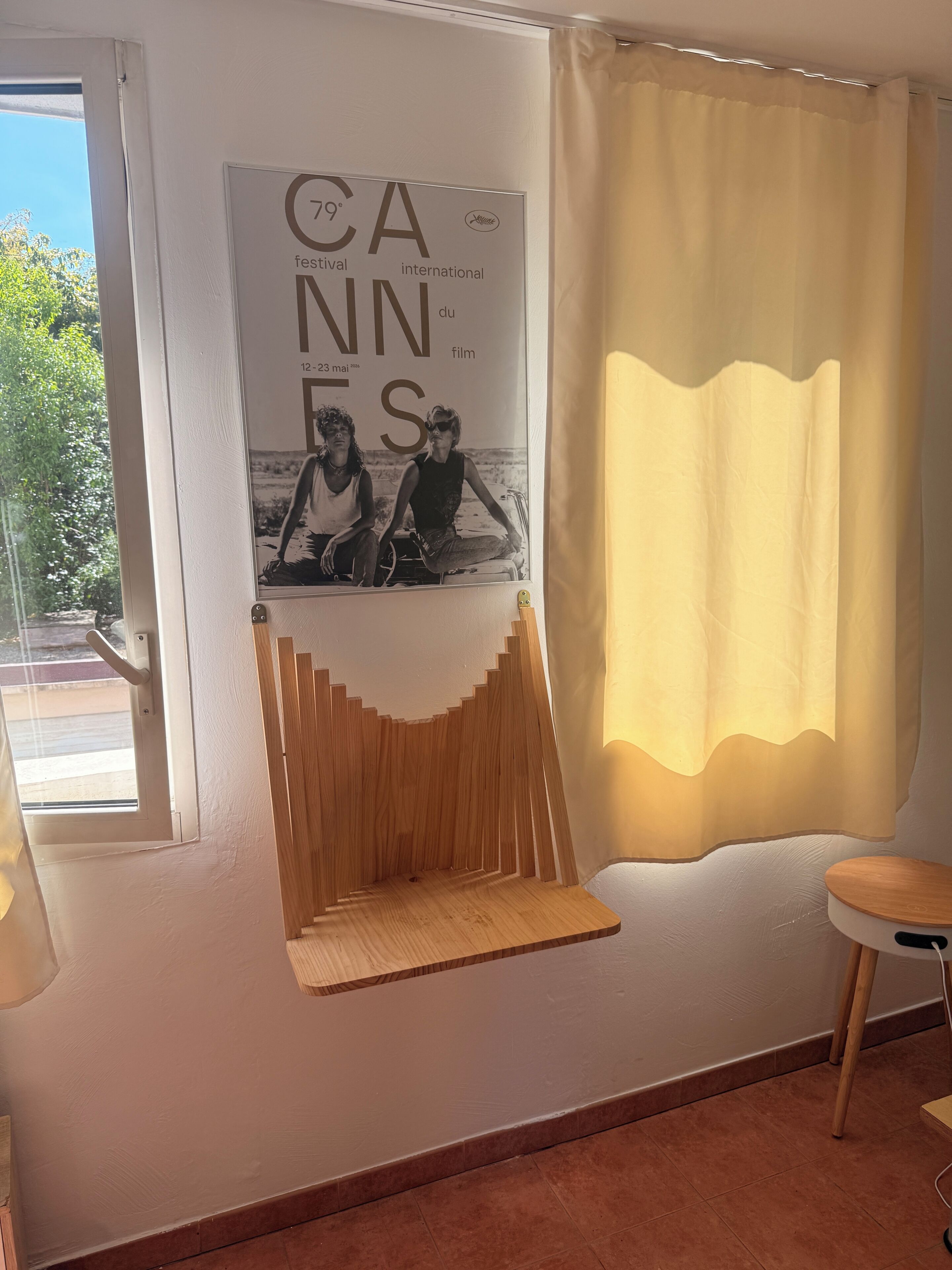 A room with a poster of the Cannes Film Festival, a window with a view of greenery, and a wooden shelf.