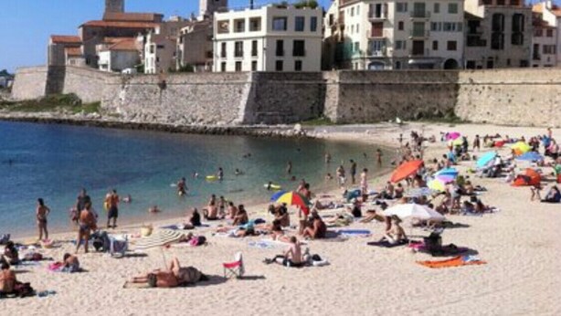 A beach with people sunbathing and swimming, a stone wall, and buildings with balconies. - Port Prestige 2 Pièces (Antibes)