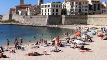 A beach with people sunbathing and swimming, a stone wall, and buildings with balconies.
