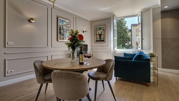 Een eetruimte met een ronde tafel, stoelen en een vaas met bloemen. Achtergrond is een bank en een radiator.