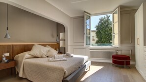 Een moderne slaapkamer met een groot bed, nachtkastjes, een rode poef en een raam met uitzicht op groen.