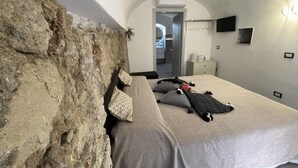 A bedroom with a stone wall, a bed with grey bedding, a flat-screen TV, and a small bedside table with a lamp. - Tufoletto Apartment (Palermo)