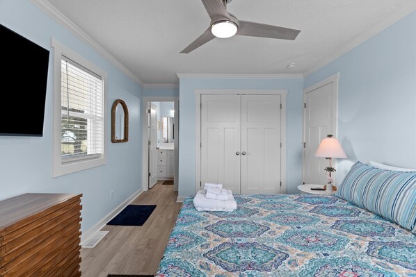 A bedroom with a bed, a ceiling fan, a dresser, a TV, and a door leading to another room.