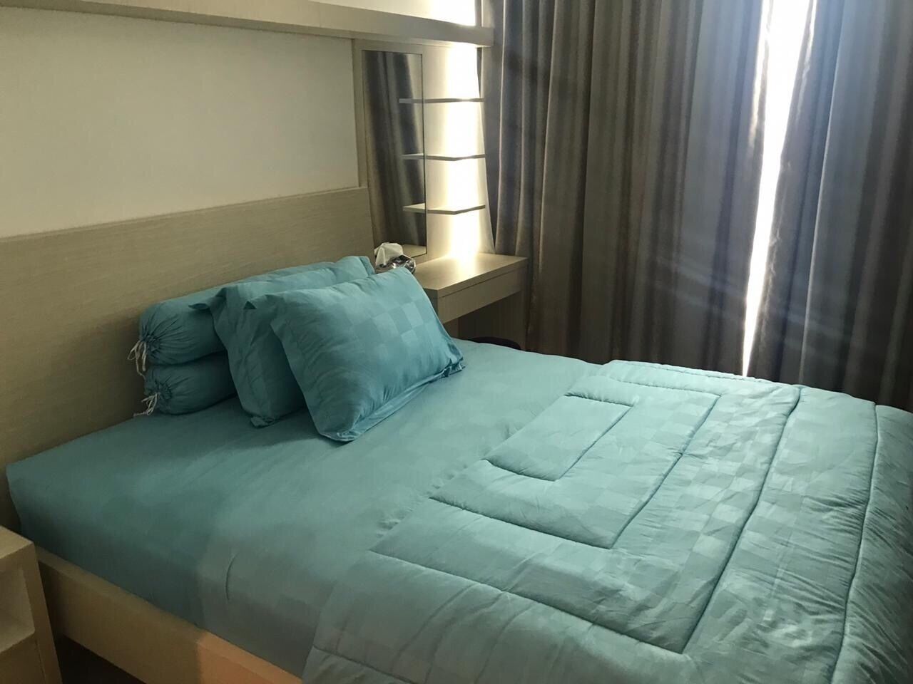 Un lit avec une couvre-lit bleu clair et des oreillers, une table de chevet, un miroir et une fenêtre avec des rideaux.