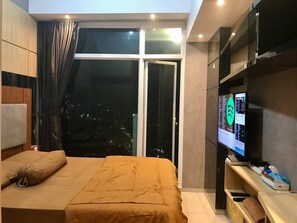 A hotel room with a bed, a TV, a window with a city view, and a sliding door. - Treepark City Apartement by Echa (Tangerang)