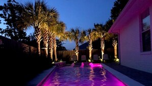A swimming pool with a purple light, surrounded by palm trees and a white building.