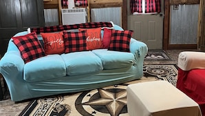 Een knus interieur met een lichtblauwe bank, een rood-zwart geruit plaid en een klein voetenbankje. Een houten plafond en een deur met raam zijn zichtbaar.