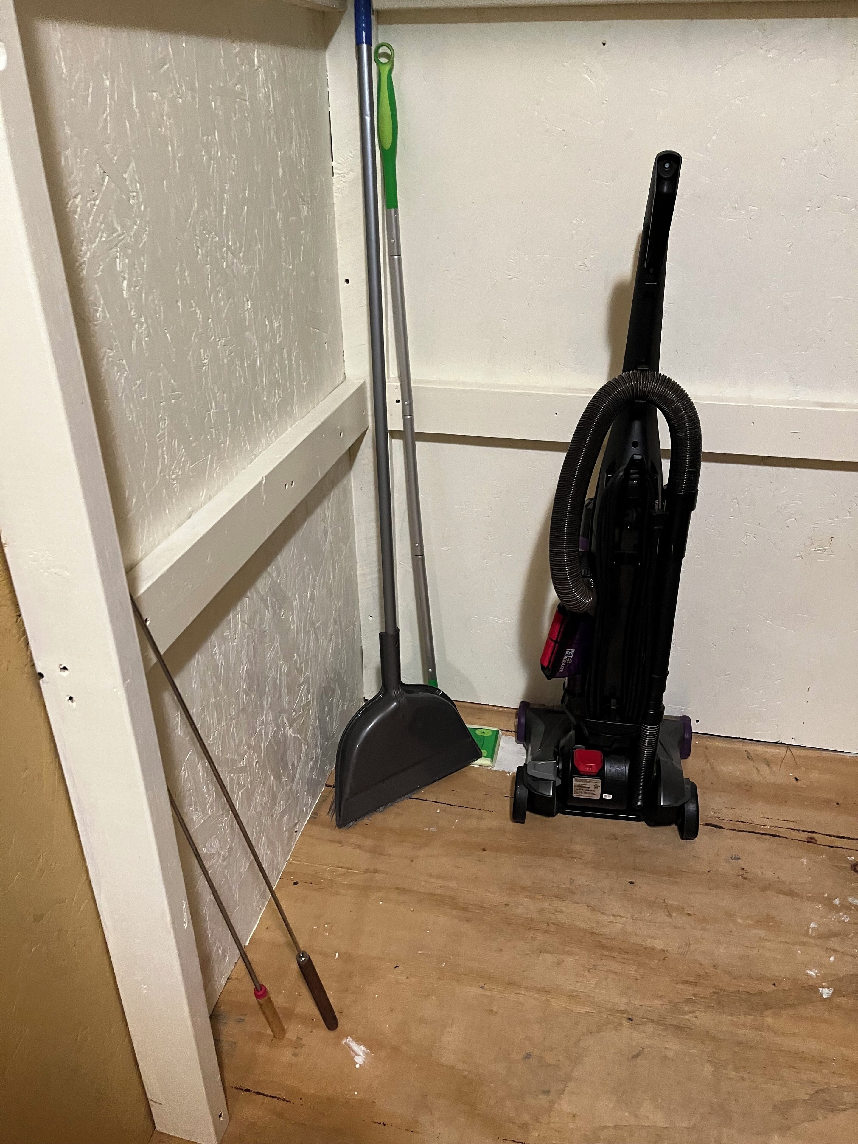 A corner of a room with a vacuum cleaner, a broom, and a dustpan leaning against the wall.