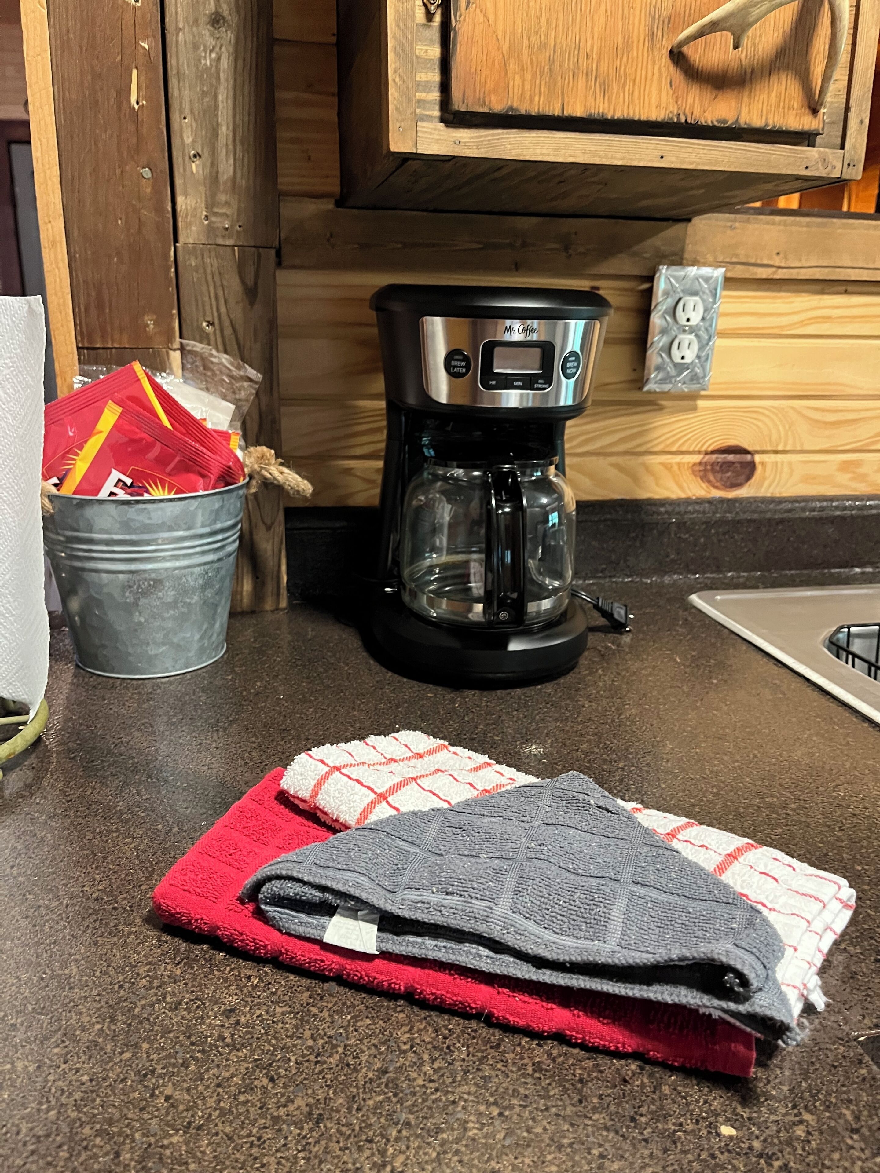 Une cuisine équipée d’une machine à café, d’un seau en métal contenant des sachets de café, et d’une serviette rouge et grise posée sur le comptoir.