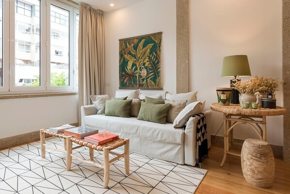 A modern living room with a white sofa, a coffee table, a patterned rug, a painting, a lamp, and a small side table with plants.