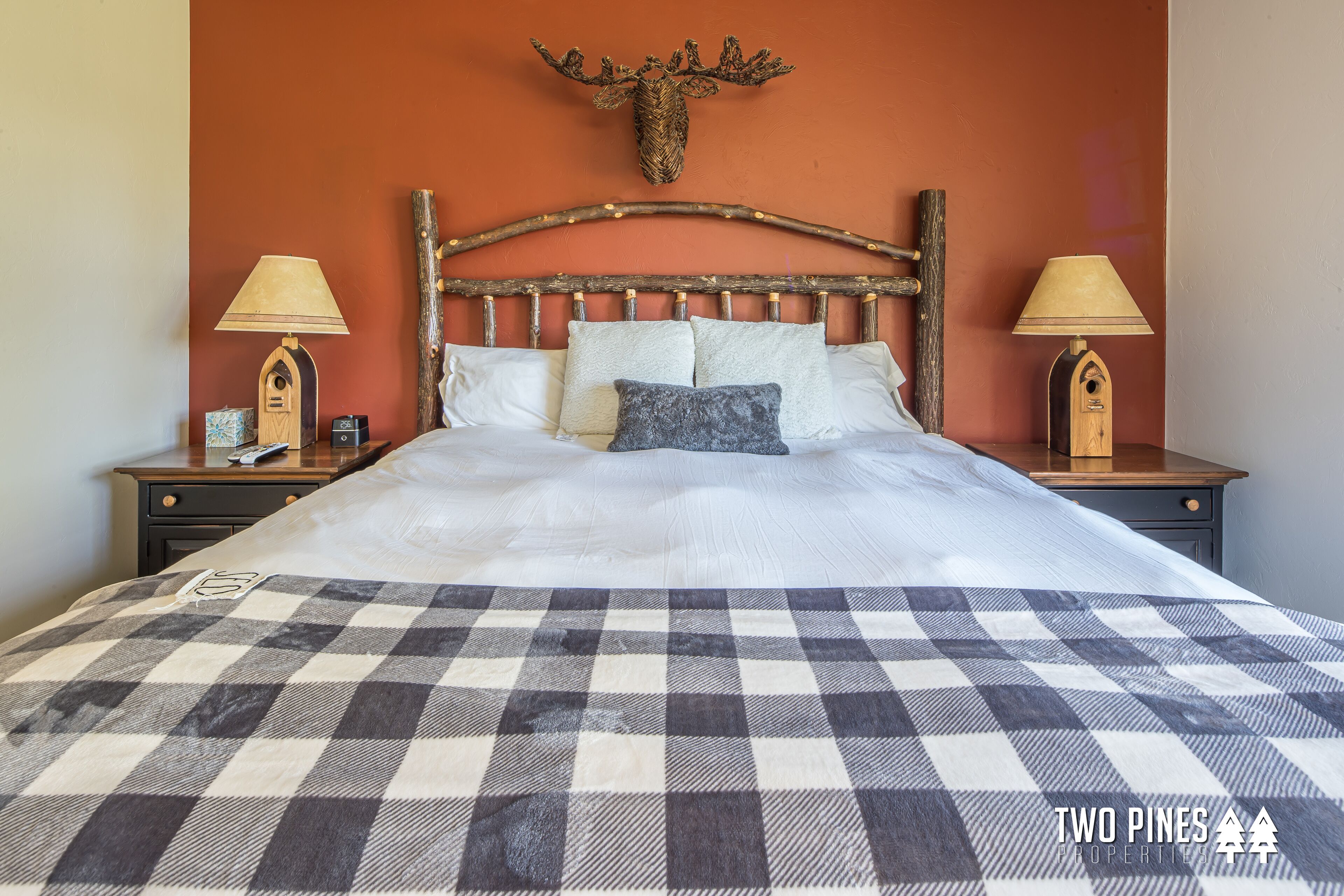 A bed with a checkered bedspread, a wooden headboard with antlers, two bedside lamps, and a nightstand with a clock and a small box.