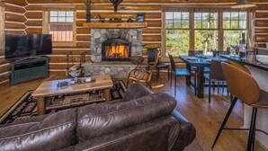A log cabin interior with a stone fireplace, leather sofa, dining table, and a television. - Pet Friendly Cabin| Hot tub, Deck & Trail (Big Sky)