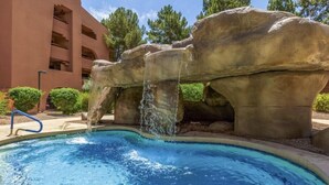 Piscina com cachoeira artificial de pedra, cercada por um edifício com varandas.