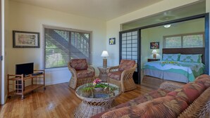 A room with a bed, two wicker chairs, a small table with a vase, a TV, and a window with blinds. - Condo 500 Feet to the Ocean (Koloa)