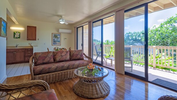 A spacious living area with a large sliding glass door leading to a deck, a wicker sofa, a glass coffee table, and a kitchen area with cabinets and a dining table. - Condo 500 Feet to the Ocean (Koloa)