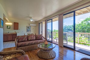 A spacious living area with a large sliding glass door leading to a deck, a wicker sofa, a glass coffee table, and a kitchen area with cabinets and a dining table. - Condo 500 Feet to the Ocean (Koloa)