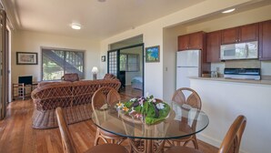 A modern kitchen with wooden cabinets, a microwave, and a stove. There is a dining area with a glass table and wicker chairs. - Condo 500 Feet to the Ocean (Koloa)