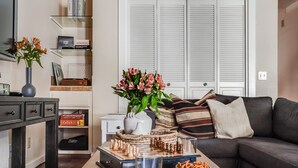 A living room with a dark-colored sofa, a coffee table with a chess set, a vase with flowers, and a bookshelf with books and decorative items.