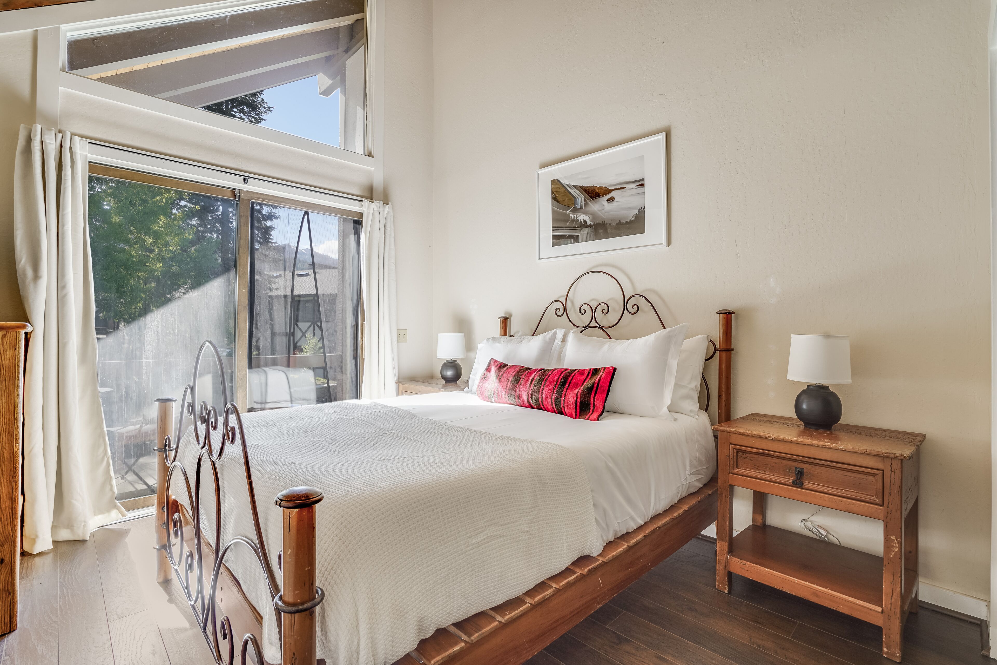 Een slaapkamer met een groot bed, een houten nachtkastje, een raam met gordijnen en een lijst met een afbeelding aan de muur.