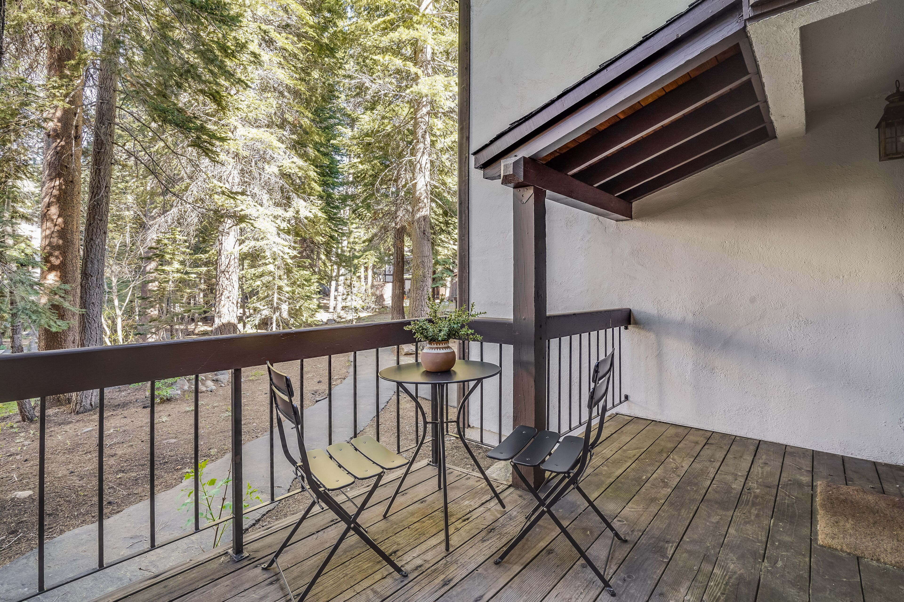 Un balcon doté d’un plancher en bois, de deux chaises, d’une petite table ronde ornée d’un pot de fleurs, et offrant une vue sur une forêt.