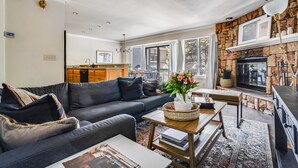 A living room with a sofa, coffee table, and fireplace. There is a kitchen in the background with wooden cabinets and a dining area.