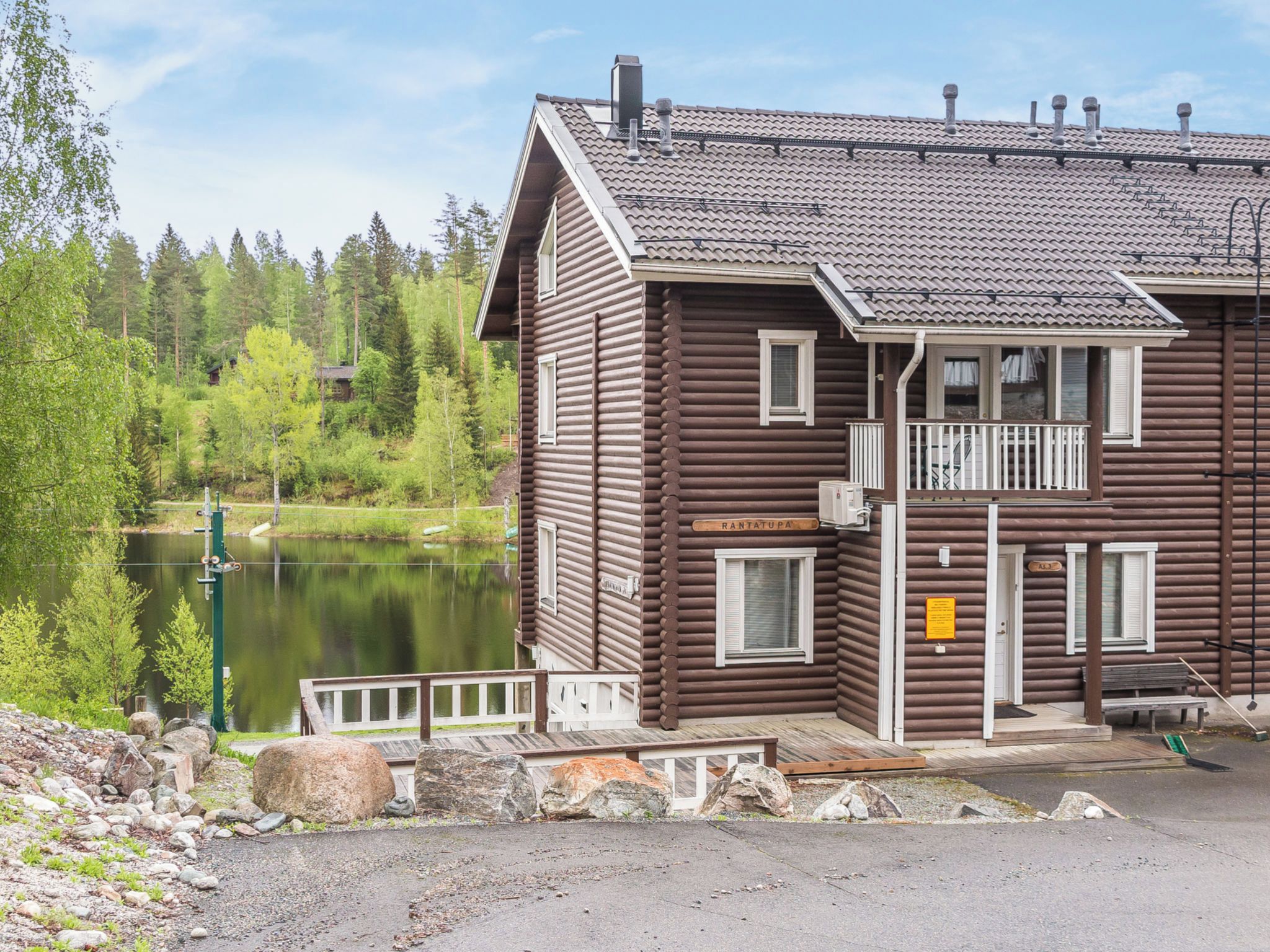 Puinen mökki, jossa on parveke, ja sen edessä on kyltti, jossa lukee ”RANTATUPA”. Mökistä avautuu näkymä järvelle.