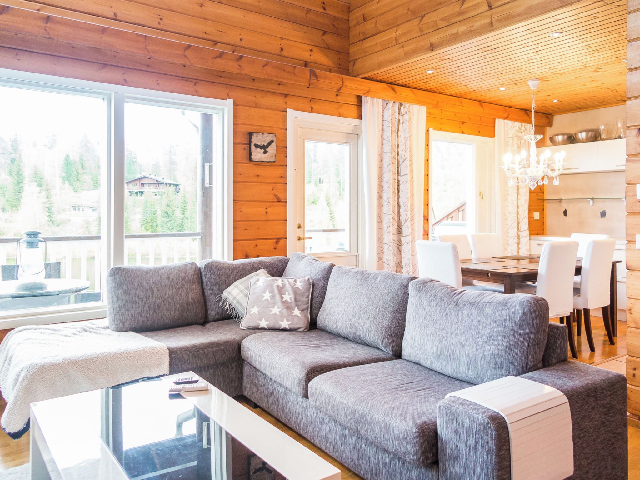 Een moderne woonkamer met een grijze bank, een glazen salontafel en uitzicht op een bos door grote ramen.