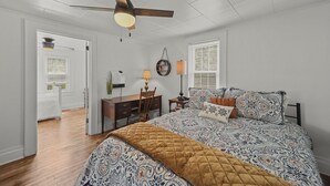 A bedroom with a large bed, a desk, a chair, a ceiling fan, and a window with blinds.