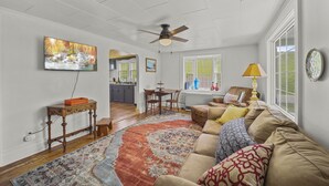 A spacious living room with a large window, a ceiling fan, a dining area, and a kitchen in the background.
