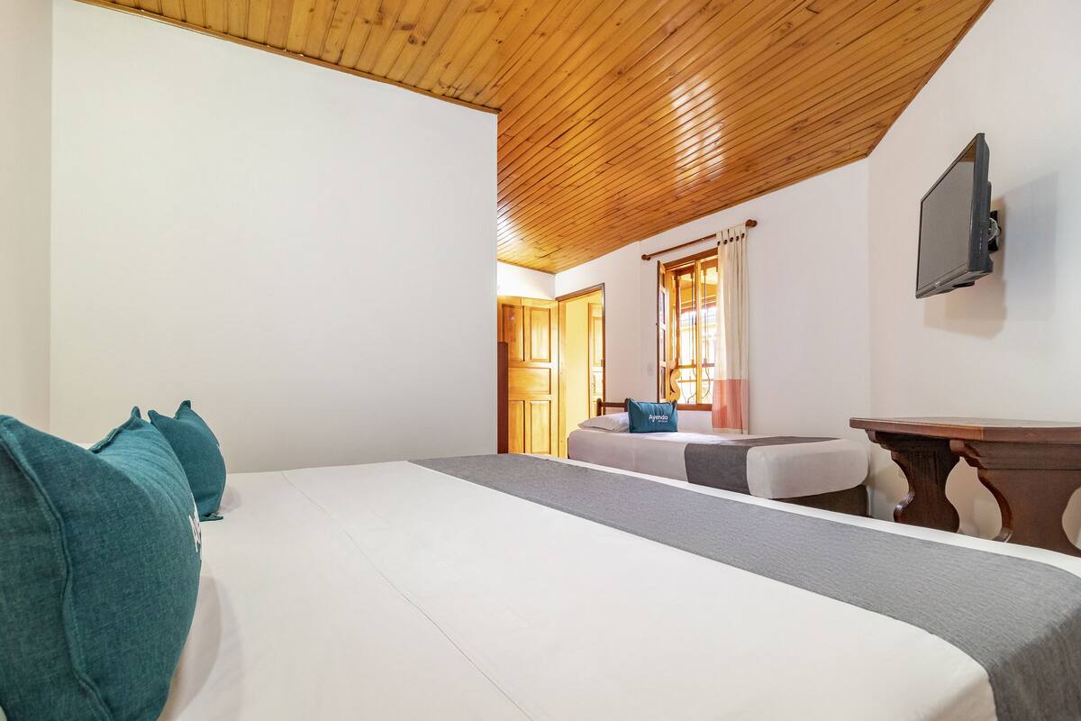 A hotel room with a wooden ceiling, a bed with teal pillows, a small table, and a TV mounted on the wall.