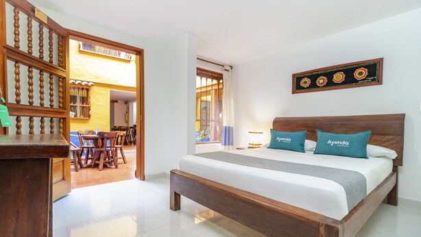 Une chambre d’hôtel avec un lit en bois, un bureau et un coin repas, visibles à travers une porte ouverte.