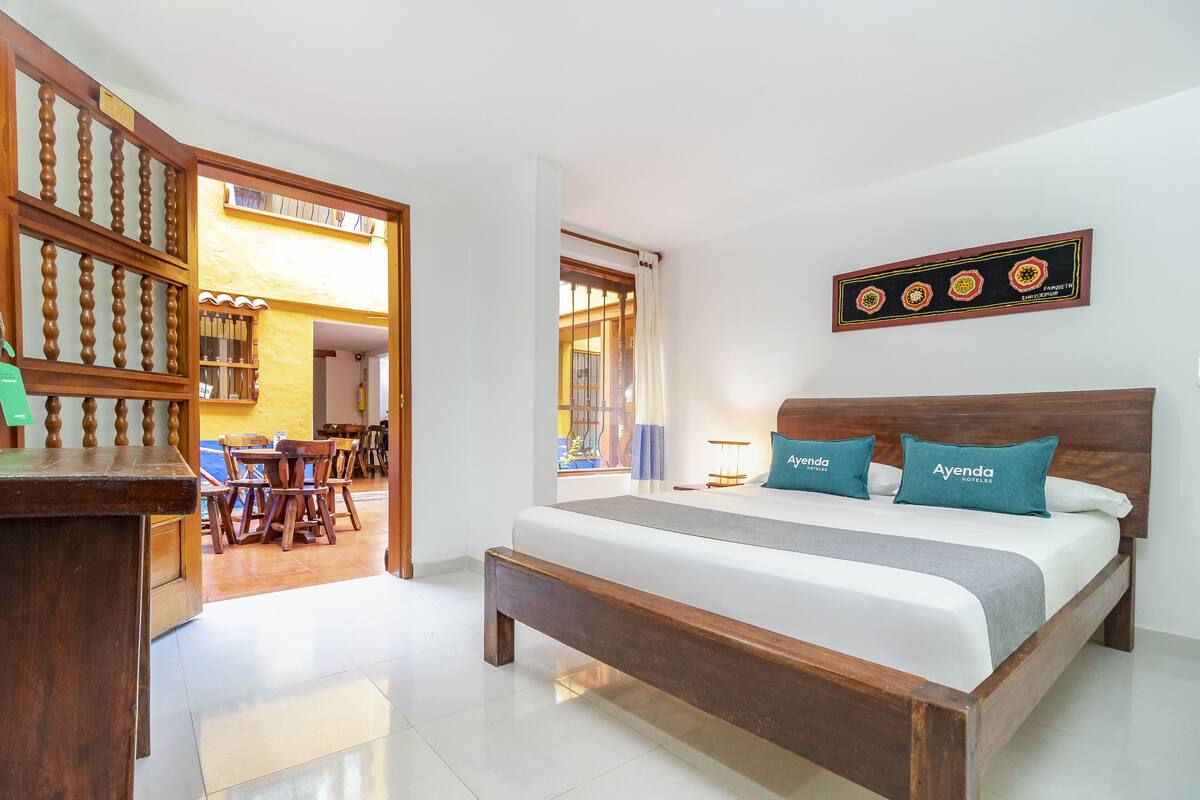 A hotel room with a wooden bed, a desk, and a dining area visible through an open door.