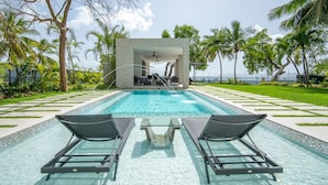 Un espace piscine doté de deux chaises longues et d’un coin salon abrité, entouré de palmiers et offrant une vue sur l’océan.