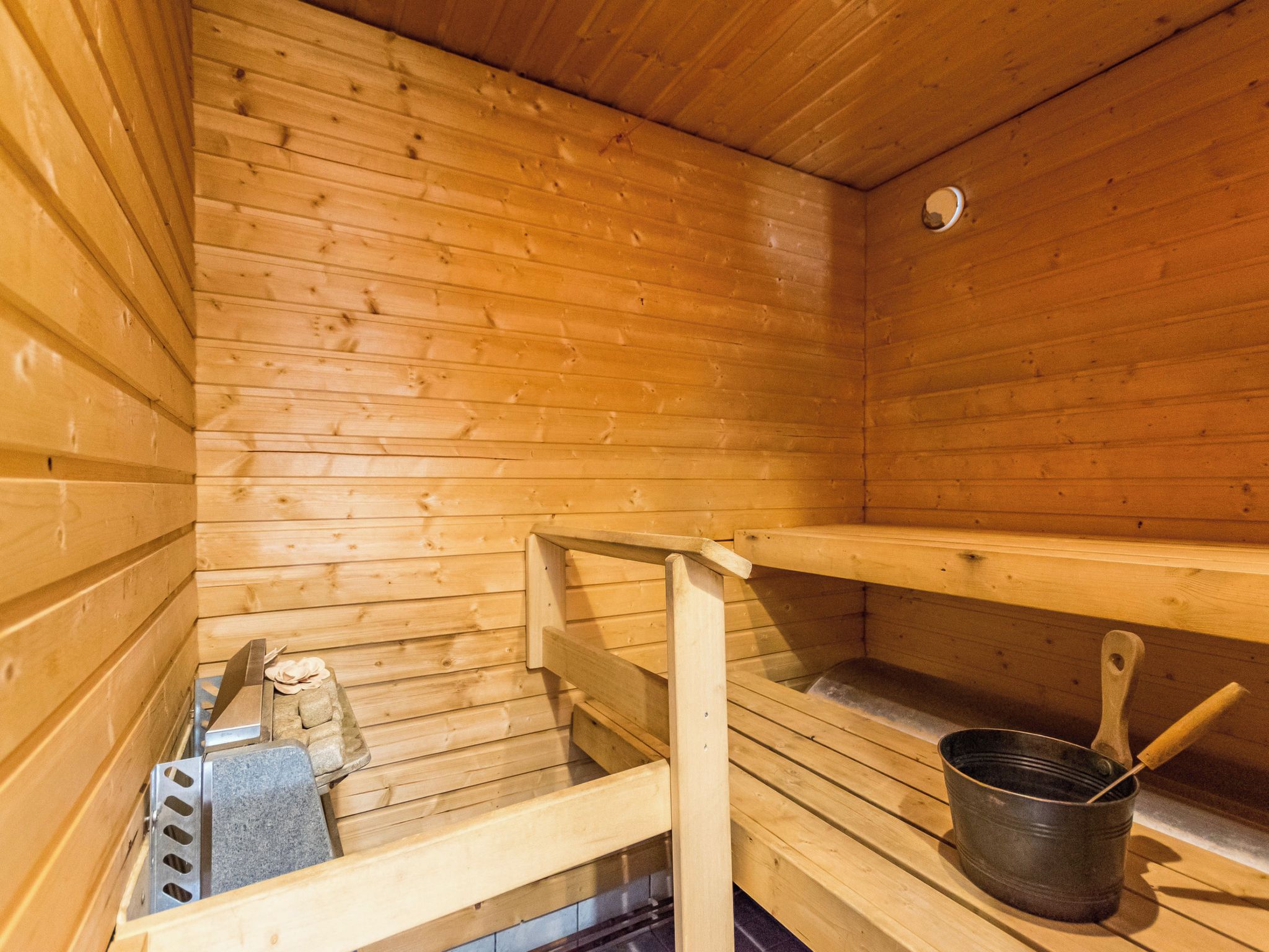 Uma sauna com paredes e bancos de madeira, uma grade de metal e um balde com cabo de madeira.