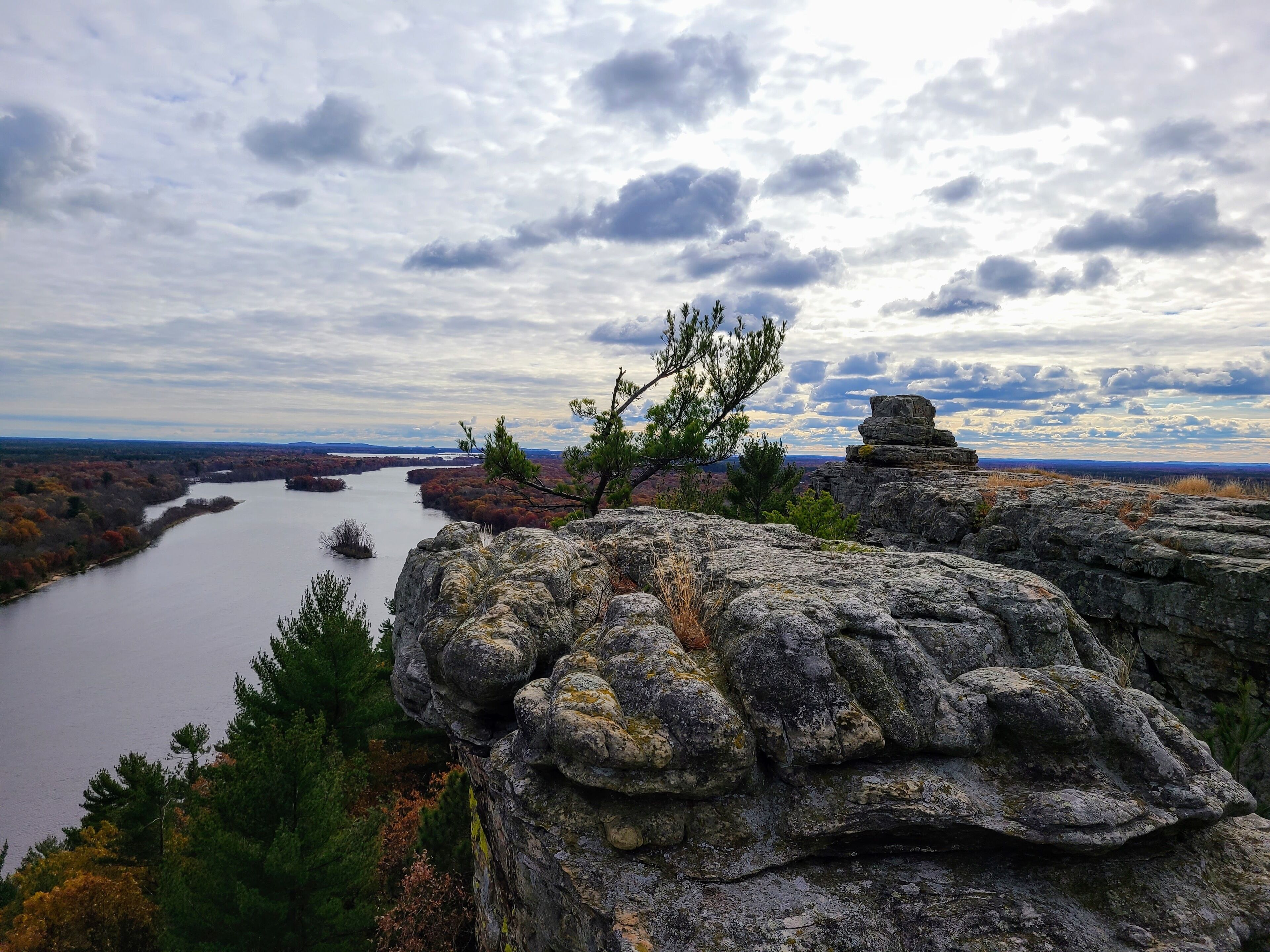 A rocky landscape with a river, trees, and a cloudy sky.