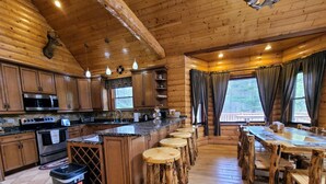 A rustic cabin-style kitchen with wooden cabinets, a central island with stools, a dining table, and a large window with curtains. - Luxury Family Cabin in a forest with a private pond (Necedah)