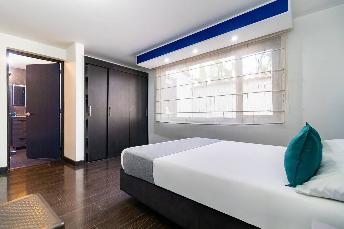 Une chambre à coucher moderne, avec un lit, une fenêtre équipée de stores, une armoire et une porte donnant sur une autre pièce.