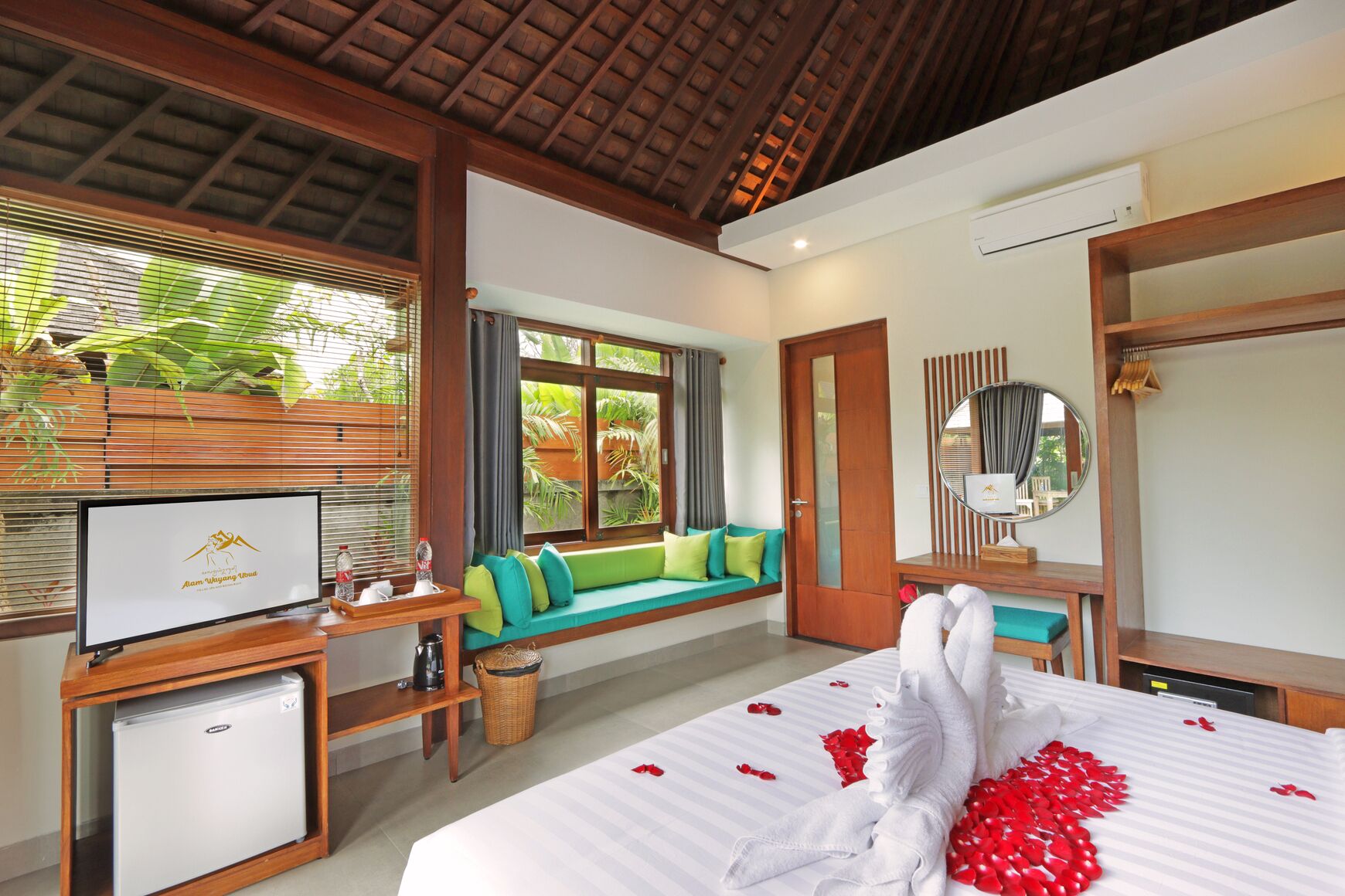 A hotel room with a bed, a TV, a small fridge, a seating area with cushions, and a window with blinds.