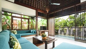 A modern living room with a blue sofa, a wooden coffee table, and a TV. Large windows provide a view of greenery outside. - Alam Wayang Ubud (Ubud)