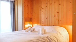 Una cama bien tendida con sábanas y toallas blancas, paneles de madera en la pared y una mesita de noche con una lámpara.