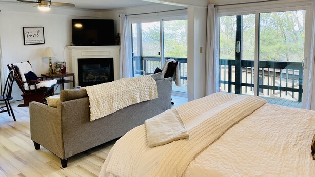 A bedroom with a large bed, a grey sofa, a fireplace, and a balcony with a view of trees. - Oz Festival! Walk to Resort! Pets! Fireplace! (Beech Mountain)