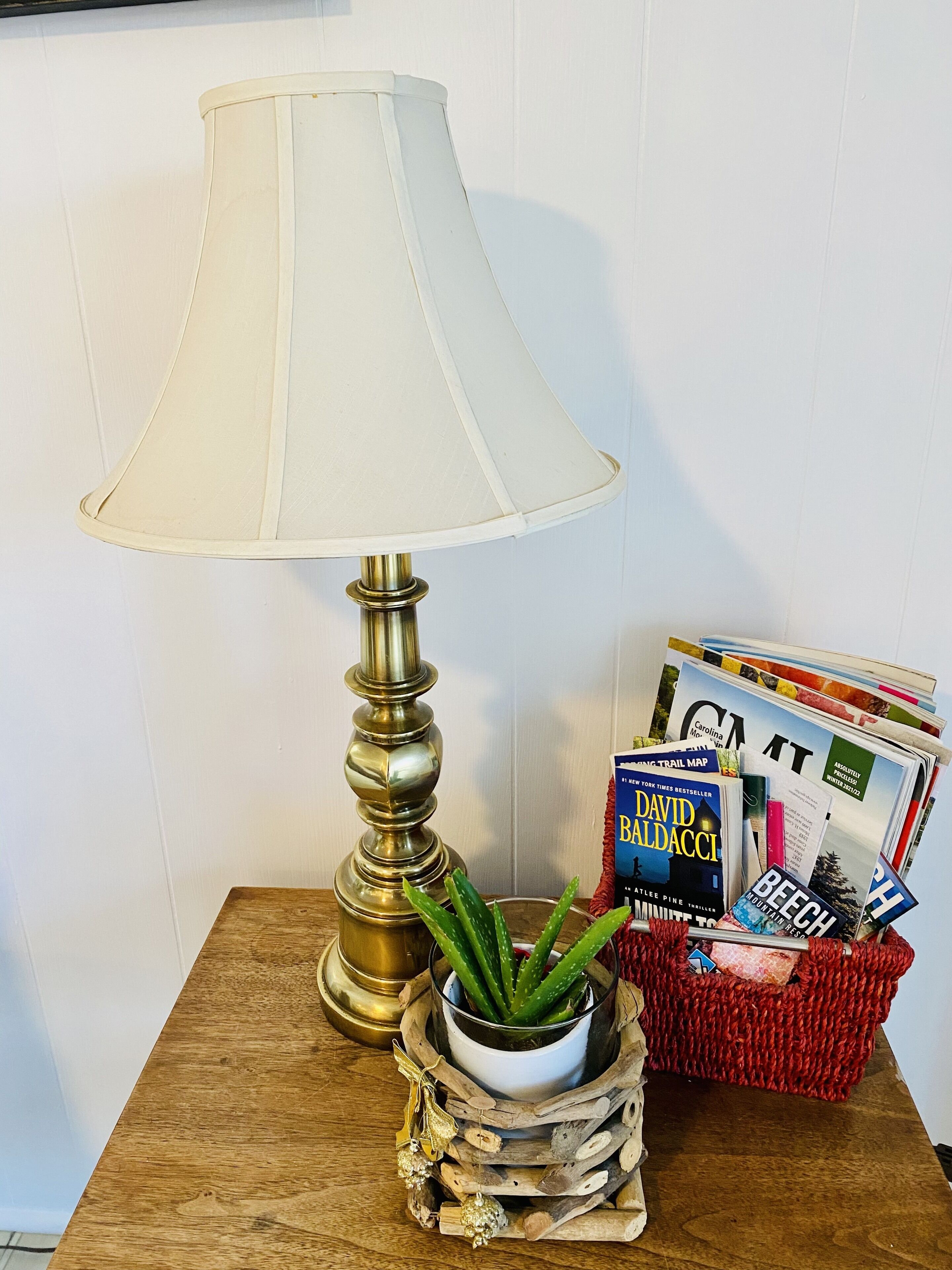 Une lampe avec un abat-jour blanc, une plante en pot et un panier rempli de magazines, le tout posé sur une table en bois.