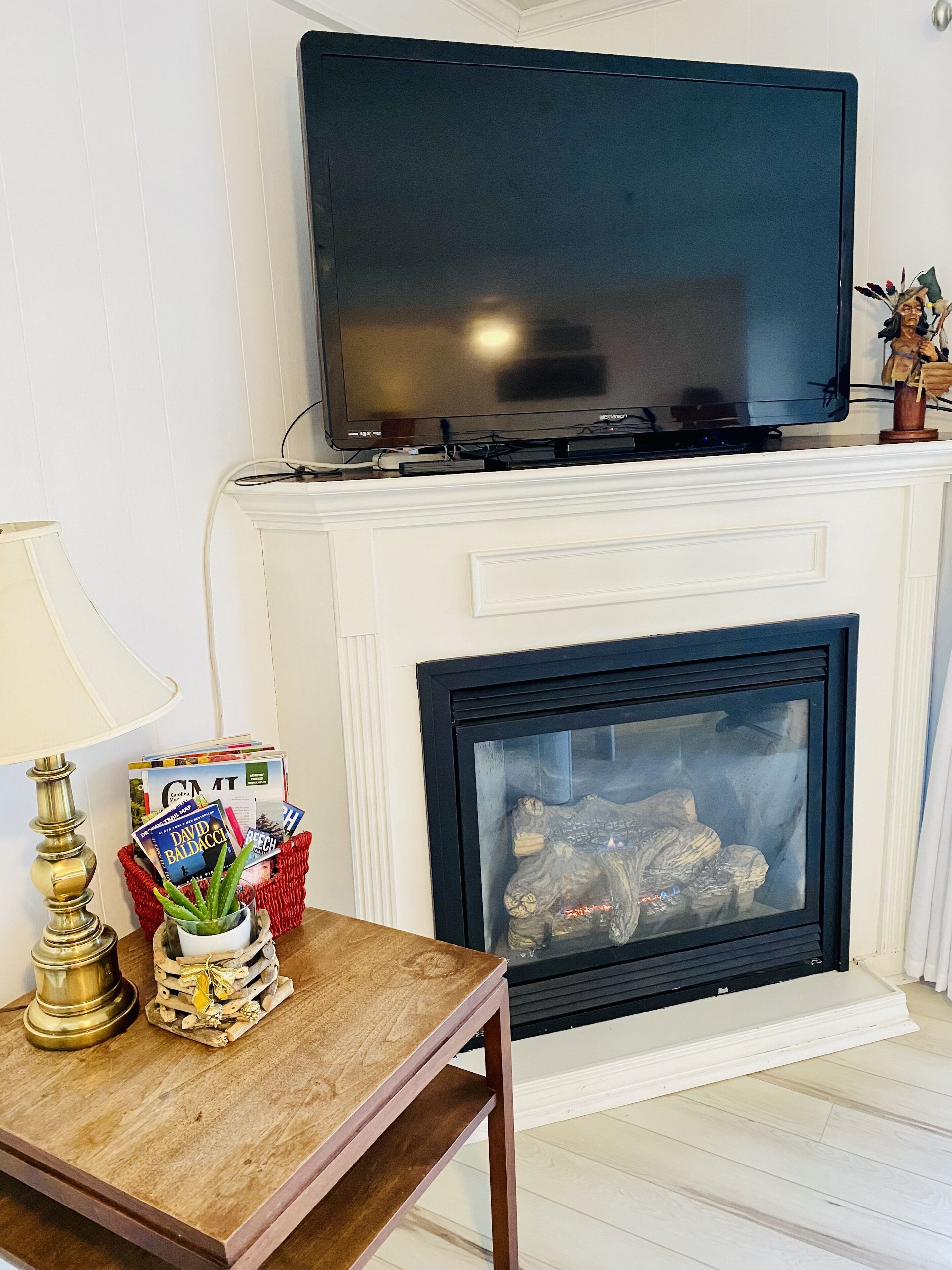 A living room with a flat-screen TV mounted above a white fireplace, a wooden table with magazines and a lamp, and a red basket with books and a plant.